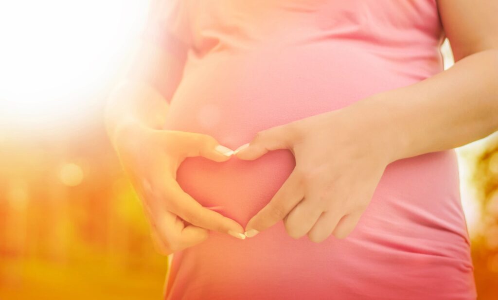 a picture of a pregnant woman wearing a customized shirt. Her fingers form a heart shape on her pregnant belly
